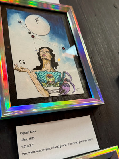 A framed art print featuring a whimsical illustration of a woman with a crab under a crescent moon, displayed on a dark wooden surface with a label card below.