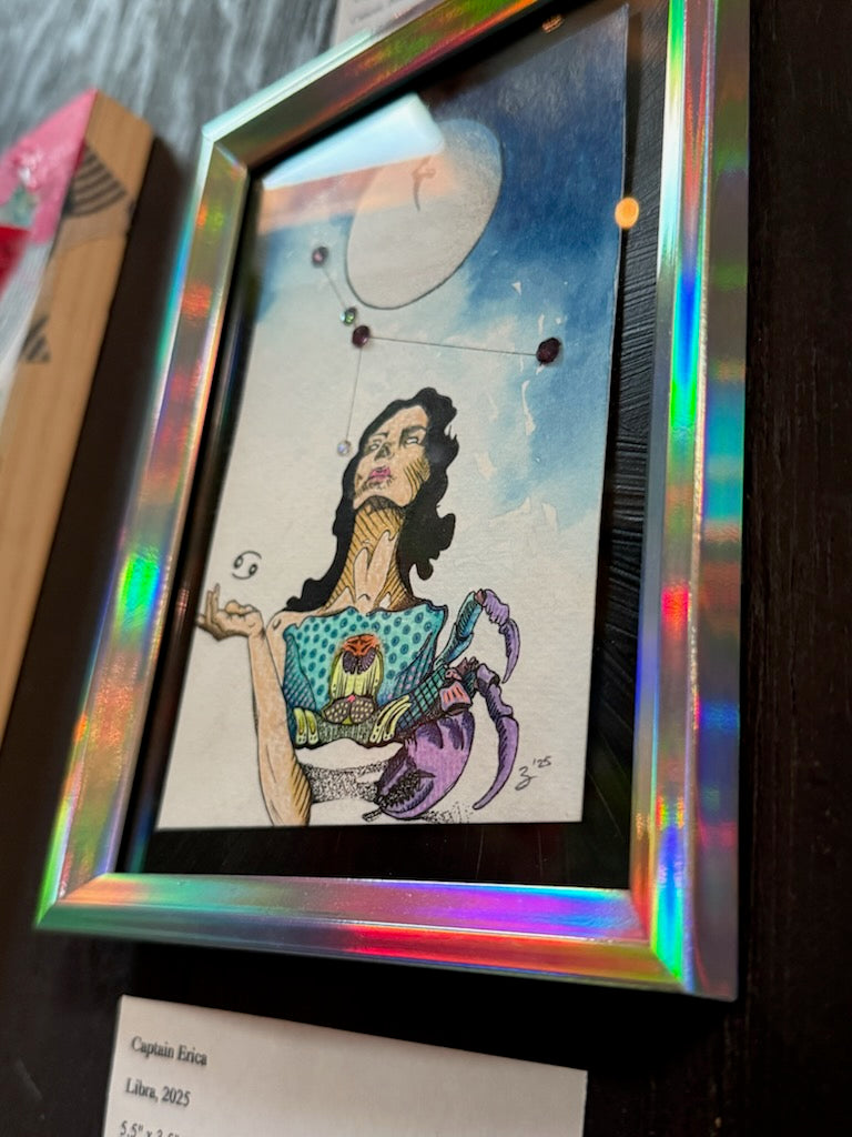 A framed art print featuring a stylized illustration of a woman with a crab, set against a blue sky with a moon and stars. The artwork is housed in a holographic iridescent frame and displayed on a dark wooden surface.