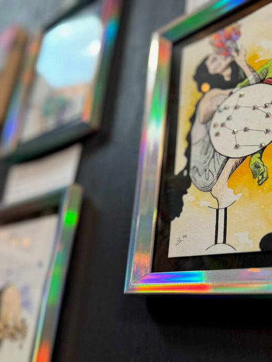 A close-up of a framed art print with an iridescent holographic frame, showing a stylized illustration of a woman holding a wine glass on a dark wall.