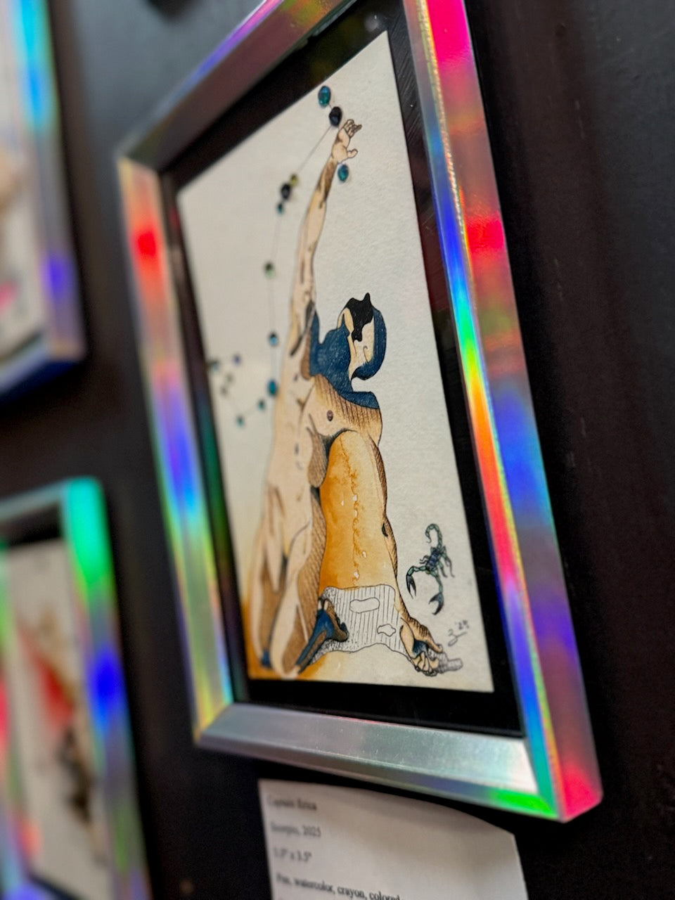 A framed art print featuring a stylized figure in beige, black, and blue tones, displayed on a dark wall with a holographic iridescent frame.