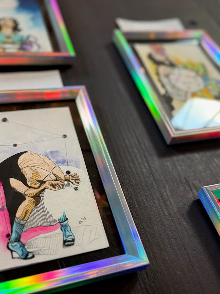 A close-up of framed art prints with an iridescent holographic frame, showing a stylized illustration of a figure in beige and blue tones. Other framed prints are visible in the background on a dark wooden surface.