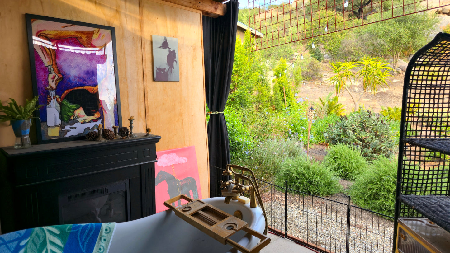 A bathroom scene with a freestanding white bathtub, brass bath tray, colorful framed wall art, a black fireplace mantel, and a large window overlooking a lush green outdoor garden.