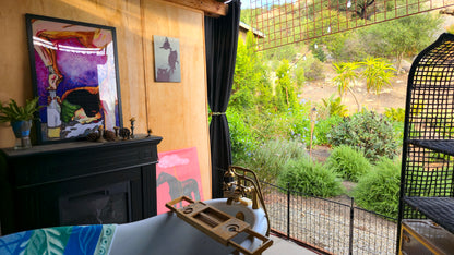 A bathroom scene with a freestanding white bathtub, brass bath tray, colorful framed wall art, a black fireplace mantel, and a large window overlooking a lush green outdoor garden.
