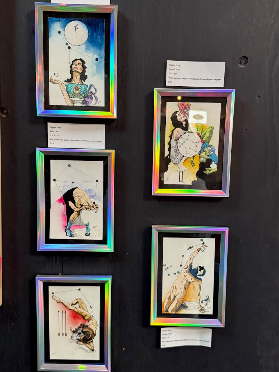 Five framed zodiac sign art prints with holographic iridescent frames displayed on a dark wall, each with a small white label card below it.